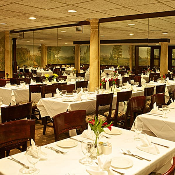Elegant Italian restaurant dining room with white tablecloths, dark wood chairs, and scenic wall murals.